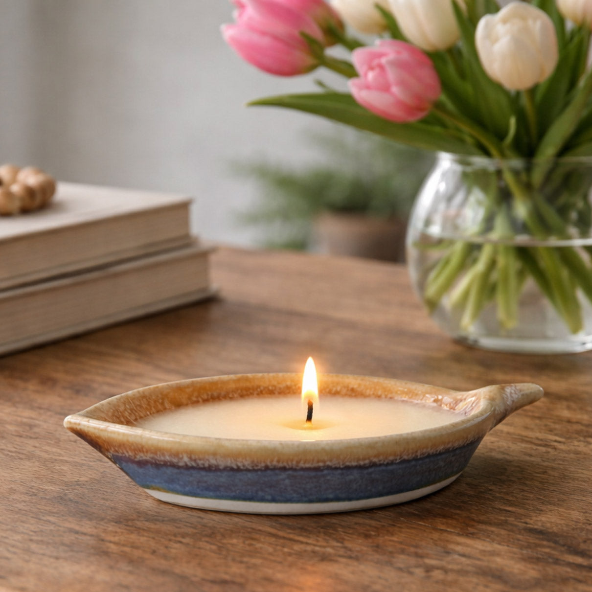 Ceramic candle holder with a lit candle on a wooden surface, next to a vase of pink and white tulips.
