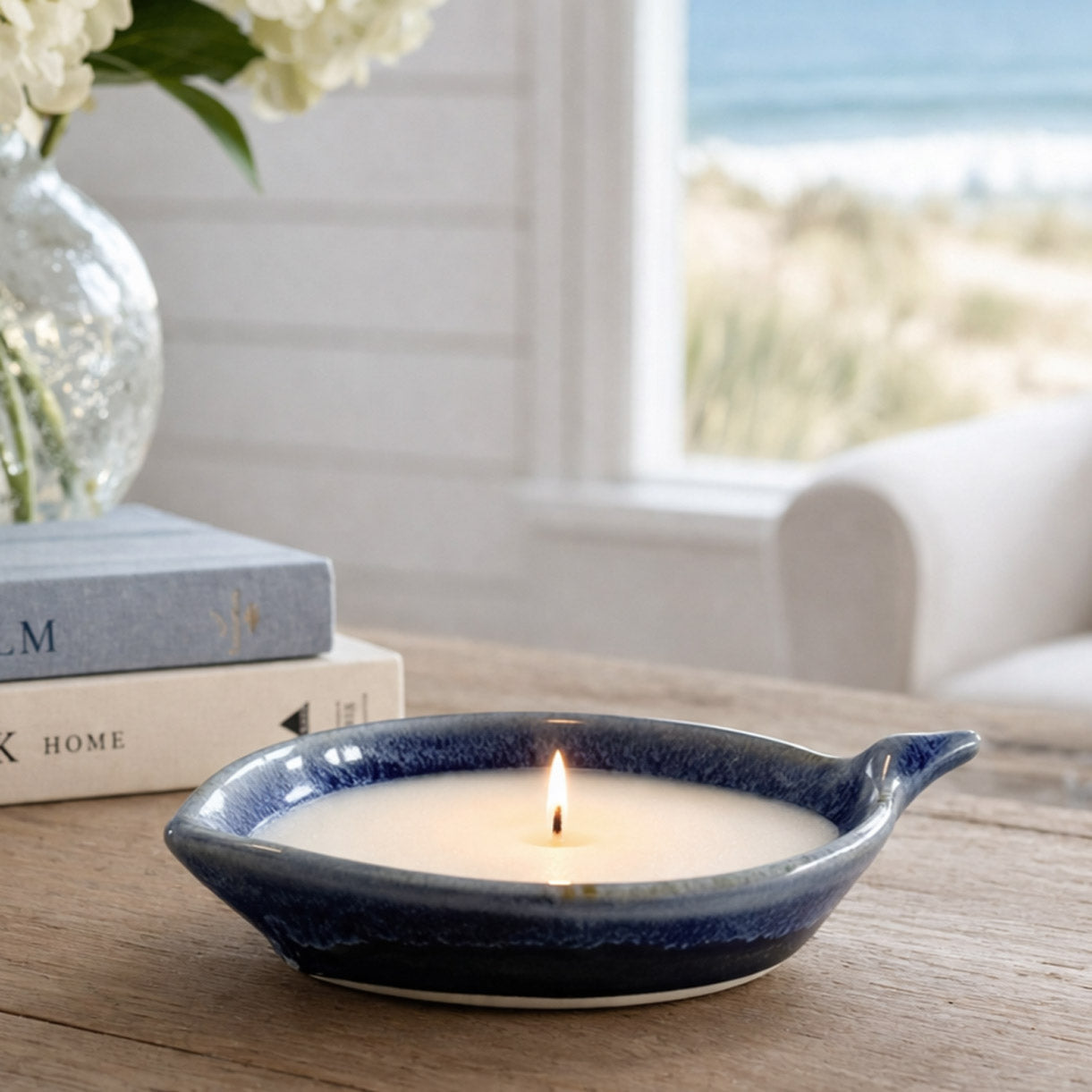 Candle in a blue ceramic holder on a wooden table with books and flowers in the background.